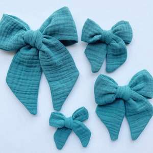 May include: Three teal blue fabric hair bows of different sizes. The bows are made of a soft, textured fabric and are tied in a classic bow shape.