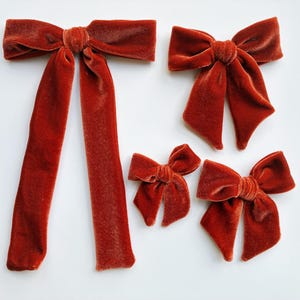 May include: Four rust-colored velvet bows of varying sizes are arranged on a white surface. The largest bow has long, flowing tails. The other bows are smaller, with classic bow shapes. The fabric has a soft, textured appearance.