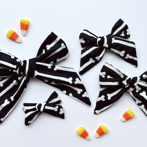 May include: Four black and white bow hair clips with a bone pattern. The bows are different sizes and are arranged on a white surface with candy corn.