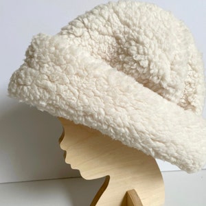 Puede incluir: Un gorro blanco y esponjoso hecho de piel sint&eacute;tica. El gorro se muestra en una cabeza de maniqu&iacute; de madera.