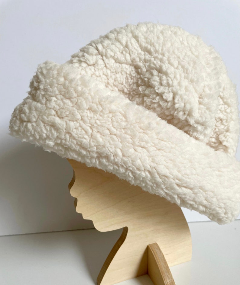 Puede incluir: Un gorro blanco y esponjoso hecho de piel sint&eacute;tica. El gorro se muestra en una cabeza de maniqu&iacute; de madera.