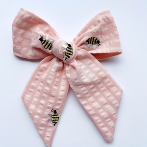 May include: A pink and white striped fabric bow with four embroidered bees. The bees are yellow and black with white wings.