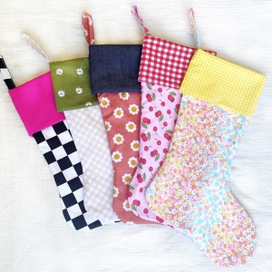 May include: Five colorful Christmas stockings with various patterns. Designs include a black and white checkerboard, white daisies on green, white daisies on a peach background, red cherries on pink, and a floral pattern. Each stocking has a different cuff and hanging loop.