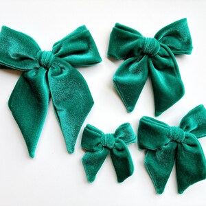May include: Five emerald green velvet bows of varying sizes are arranged on a white surface. The bows have a soft, textured appearance and are tied in the center. The bows are a rich, jewel-toned green.