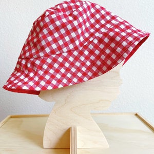 May include: Red and white gingham bucket hat. The hat is made of cotton fabric and has a wide brim.