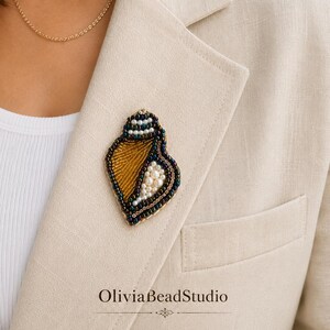 May include: A seashell-shaped brooch crafted with intricate beadwork, featuring a golden-brown centre, white pearl accents, and a dark beaded border. The piece is pinned to a beige blazer, with the text OliviaBeadStudio printed below the brooch.