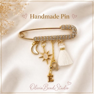May include: A gold-tone safety pin brooch featuring a crystal-encrusted bar, a white crescent moon charm, a sun charm with a white center, a white tassel, a crystal-studded letter E, and a small gold-tone charm. Text reads Handmade Pin and Olivia Bead Studio.