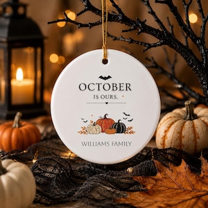 Op de afbeelding: Een rond wit keramieken ornament dat aan een gouden koord hangt. De tekst luidt OCTOBER IS OURS. en WILLIAMS FAMILY. Het ontwerp bevat een zwarte vleermuis, een klein hartje en een groepje oranje, zwarte en witte pompoenen met herfstbladeren.