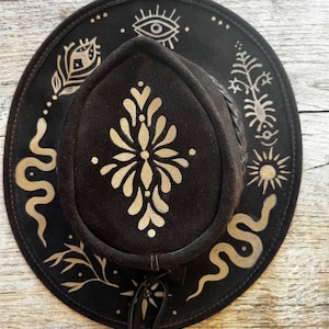 Könnte beinhalten: Ein dunkelbrauner Wildlederhut mit goldfarbenen, aufgemalten Motiven, darunter ein zentrales florales Muster auf der Krone sowie Symbole auf der Krempe wie Schlangen, ein Auge, eine Sonne und botanische Elemente mit einem geflochtenen Hutband.