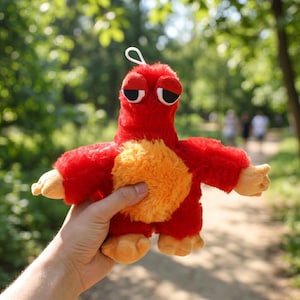 Puede incluir: Un peluche rojo con forma de criatura con vientre naranja y manos y pies color beige. Tiene un lazo blanco en la parte superior. El juguete se sostiene en una mano sobre un fondo verde borroso.