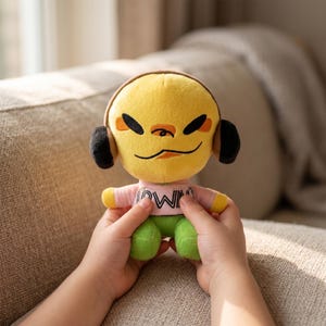 May include: A plush doll with a yellow head, black headphones, and a pink shirt with the word "OWN" printed on it. The doll has green trousers and is held by a person. The doll's face has black eyes and a small orange nose.