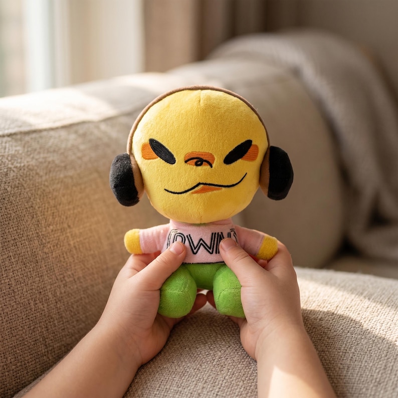 May include: A plush doll with a yellow head, black headphones, and a pink shirt with the word "OWN" printed on it. The doll has green trousers and is held by a person. The doll's face has black eyes and a small orange nose.