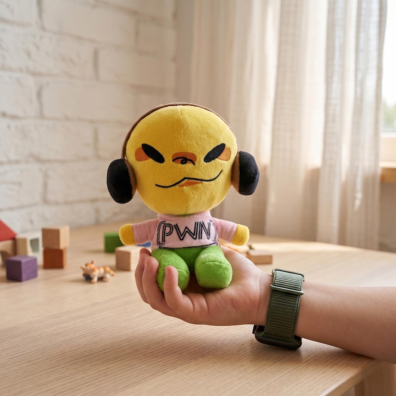 May include: A yellow plush toy with a smiling face, black headphones, and green trousers. The toy is wearing a pink t-shirt with the letters "PWN" printed on it. The toy is held in a person's hand.