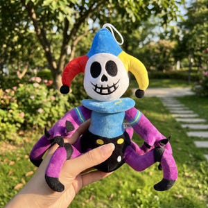 May include: A plush toy in the shape of a jester with a skull-like face. The jester has a blue, red, and yellow hat, purple arms and legs, and a blue collar. The toy is held in a person's hand.