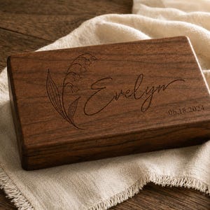 Puede incluir: Una caja rectangular de madera marrón oscuro con tapa abatible. La superficie superior presenta un diseño grabado con láser de una flor de lirio de los valles, el nombre Evelyn en cursiva y la fecha 06.18.2024 grabada a la derecha.
