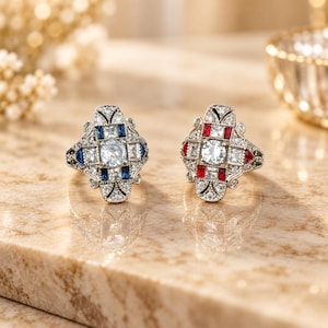 Puede incluir: Dos anillos de tono plateado con intrincado trabajo en metal y piedras centrales redondas transparentes. Un anillo está acentuado con piedras azules rectangulares, mientras que el otro presenta piedras rojas rectangulares. Ambos están rodeados de pequeñas piedras transparentes.