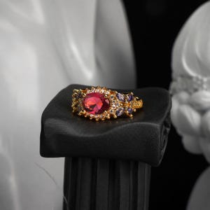 Puede incluir: Un anillo de tono dorado con una gran piedra central roja de corte ovalado rodeada por un halo de pequeñas piedras blancas transparentes. La banda está decorada con piedras moradas de corte marquesa y piedras blancas.