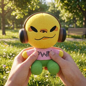 May include: A yellow plush toy with black eyes, brown headphones, a pink shirt with white text, and green trousers. The toy is held in front of a blurred park background with green grass and trees.