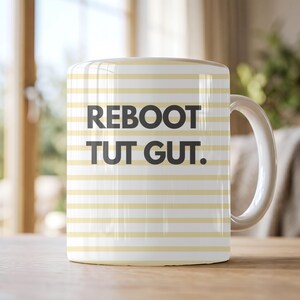 Könnte beinhalten: Ein weißer Keramikbecher mit horizontalen hellgelben Streifen und dem schwarzen Text REBOOT TUT GUT. mittig auf der Vorderseite. Der Becher hat einen geschwungenen Henkel auf der rechten Seite und einen glatten, zylindrischen Körper.