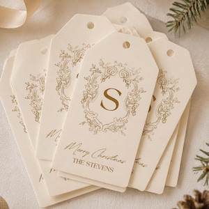 Op de afbeelding: Crèmekleurige cadeaulabels met gouden letters en een decoratief kransontwerp. De labels tonen de letter "S" en de tekst "Merry Christmas" en "The Stevens". Een feestelijk vakantiethema.