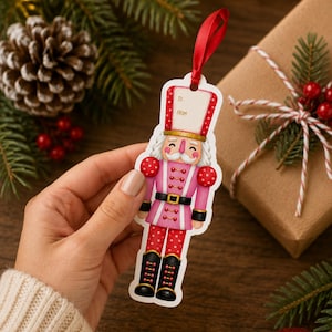 May include: A die-cut gift tag shaped like a nutcracker, featuring a pink and red color palette with white polka dots. The nutcracker wears a tall hat with a blank 'TO' and 'FROM' section, and a red satin ribbon is attached to the top for hanging.