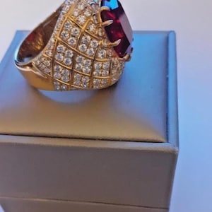 May include: A gold ring with a large, rectangular, deep red gemstone. The ring is encrusted with numerous small, clear gemstones, set in a grid pattern. The ring is displayed on a gray jewelry box.