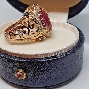 May include: A gold ring with a large, oval, red gemstone, surrounded by smaller diamonds and set in an ornate filigree design. The ring is displayed in a dark blue jewelry box with a cream-colored interior.
