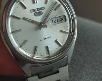 Peut inclure: Montre-bracelet automatique Seiko 5 argentée avec un cadran blanc. Le cadran affiche le jour et la date, avec "TUE 26" visible. Le bracelet est en métal argenté. Le mot "AUTOMATIC" est imprimé sur le cadran. Design classique.