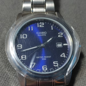 May include: A silver Casio Quartz watch with a blue dial. The watch has a date window at the 3 o'clock position and is water-resistant to 50m. The watch features a silver band and a clear protective cover.