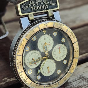 Puede incluir: Reloj de pulsera Camel Trophy vintage con esfera oscura, bisel dorado y correa de cuero marrón. El reloj presenta múltiples esferas secundarias, zonas horarias y el logotipo Camel Trophy. La caja del reloj mide aproximadamente 4 cm de diámetro.