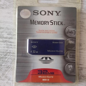 Pode incluir: Um cartão de memória Sony Memory Stick azul com 32MB de capacidade e tecnologia MagicGate, dentro da embalagem original de plástico transparente. A embalagem apresenta o logótipo Sony, o texto Memory Stick e um gráfico vermelho 32MB MSH-32.