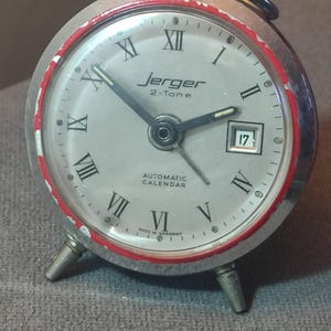 May include: A vintage Jerger 2-Tone alarm clock with a silver face, black hands, and Roman numeral hour markers. The clock has a red accent ring and a date display showing the number 17. The words "AUTOMATIC CALENDAR" and "MADE IN GERMANY" are visible.