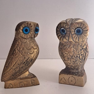 May include: Two brass owl figurines with textured feather details and bright blue circular eyes. Each owl stands on a rectangular base featuring the engraved Greek letters Alpha, Omicron, and Epsilon, reading AOE.