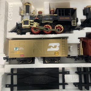Pode incluir: Um conjunto de comboio modelo com uma locomotiva preta e dourada com rodas vermelhas, um vagão de carga bege com um grande logótipo "B" branco e um vagão vermelho. O conjunto inclui peças de pista e é embalado numa caixa branca.