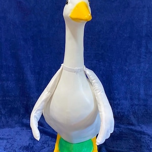 May include: A white goose figurine with a bright yellow beak and orange feet. The goose features a long neck, black eyes, and a white fabric cape attached around the neck with a thin white elastic band. The base of the figurine is green.
