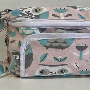 May include: A rectangular, zippered bag with a light pink owl pattern, featuring owls in teal and gray. The bag has a white trim, a front pocket, and a matching strap. The bag is designed for carrying items.
