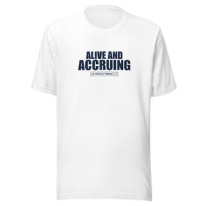 Op de afbeelding: Een wit T-shirt met korte mouwen en de donkerblauwe tekst ALIVE AND ACCRUING in het midden op de borst. Onder de hoofdtekst staat een kleiner rechthoekig kader met de code-achtige tekst =STATUS(TODAY()). Het T-shirt heeft een ronde hals.