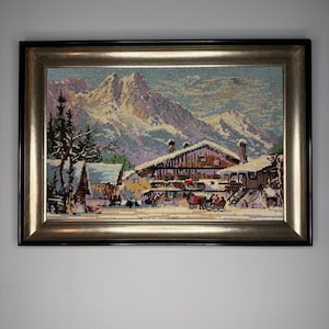 Könnte beinhalten: Gerahmtes Gobelin-Kunstwerk, das eine verschneite Bergdorf-Szene darstellt. Das Kunstwerk zeigt eine detaillierte Landschaft mit schneebedeckten Gebäuden, Bäumen und Bergen im Hintergrund. Der Rahmen ist schwarz mit einem goldenen Innenrand.