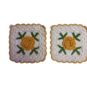 May include: Two square crochet coasters featuring a white base with a central yellow rose motif surrounded by green leaves. The edges are finished with a scalloped border in a golden yellow yarn, creating a textured and detailed floral design.