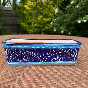 May include: A rectangular ceramic planter with a glossy dark blue glaze, featuring hand-painted white floral motifs and light blue horizontal stripes along the rim and base. The interior is finished in a smooth white glaze with a terracotta base.