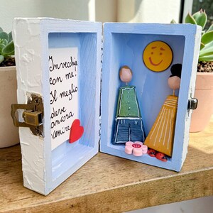 Può includere: Una piccola scatola di legno aperta, con interno blu ed esterno bianco. All'interno, una coppia dipinta è in piedi vicino a una faccina gialla sorridente. Una nota con testo italiano e un cuore rosso sono visibili. La scatola è su una superficie di legno.