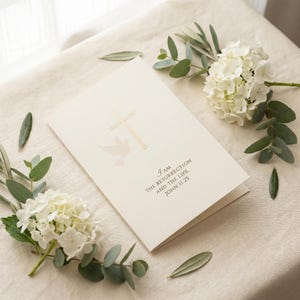 May include: A cream-colored card with a gold cross and dove design, with the text "I AM THE RESURRECTION AND THE LIFE JOHN 11:25". The card is surrounded by white flowers and green leaves.
