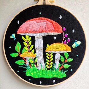 May include: Embroidered hoop art featuring three mushrooms on a black background. The largest mushroom has a red cap and white stem, while the two smaller mushrooms have yellow caps and white stems. The mushrooms are surrounded by green leaves and a few white stars.