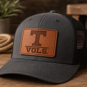 May include: A charcoal grey trucker hat with a black mesh back. The hat features a rectangular brown leather patch with the letter "T" and the word "VOLS" in orange.