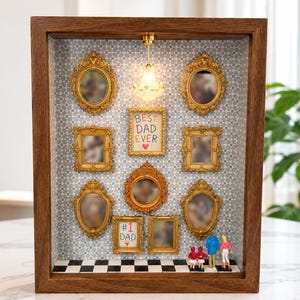 Puede incluir: Una caja de sombra de madera con una lámpara de araña en miniatura, un suelo de cuadros blancos y negros y varios marcos dorados ornamentados. Los marcos contienen imágenes borrosas y dos letreros que dicen BEST DAD EVER y #1 DAD con corazones rojos.