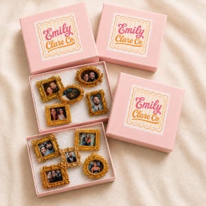 May include: Two light pink gift boxes with Emily Clare Co. labels, each containing several small, ornate gold-colored picture frames. The frames are rectangular, oval, and circular, featuring various photographs of people and a cat inside them.