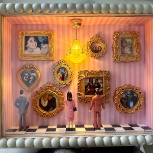 May include: A shadow box display with a white beaded frame, featuring miniature framed photos and figurines. The interior has pink and white striped wallpaper, a checkered floor, and a small chandelier. The photos are in ornate gold frames.