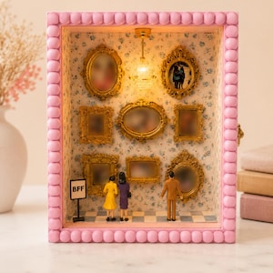 May include: A miniature diorama box with a pink beaded frame, featuring a floral wallpaper interior, a small ceiling light, and several ornate gold picture frames on the wall. Three miniature figures stand on a checkered floor near a sign labeled BFF.