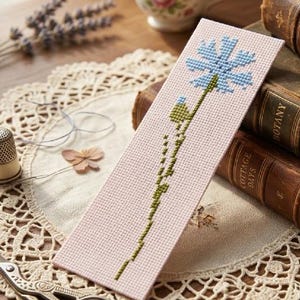 Può includere: Segnalibro fatto a mano a punto croce con un motivo floreale blu su sfondo rosa. L'immagine presenta libri antichi, una tazza da tè e accessori da cucito, evocando la lettura e l'artigianato.