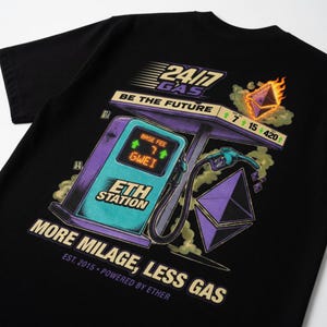 Puede incluir: Camiseta negra con un gráfico de una gasolinera con la etiqueta "ETH STATION". El diseño incluye el texto "24/7 GAS BE THE FUTURE" y "MORE MILAGE, LESS GAS". La camiseta presenta una combinación de colores turquesa y morado con una estética futurista.